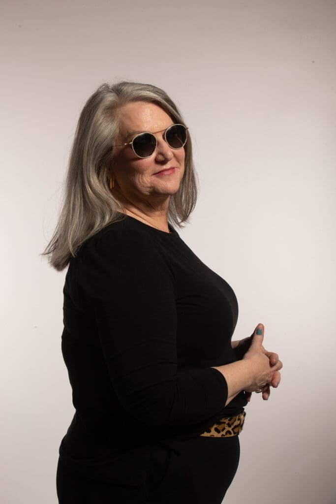 Woman wearing dark outfit and sunglasses, turned to the left and looking over her shoulder