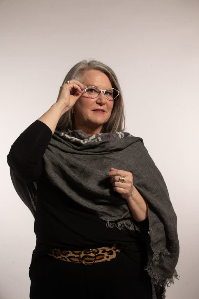 Woman wearing dark outfit and silver glasses, with one hand touching the side of her frames.
